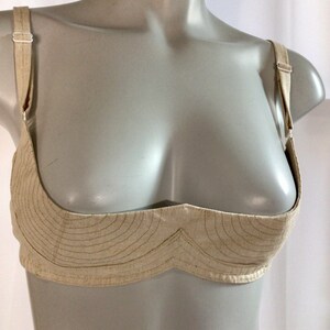 May include: A beige linen bra with adjustable straps. The bra has a unique design with a curved, scalloped neckline and a decorative stitch pattern. The text "Linen Bra" is printed below the bra.