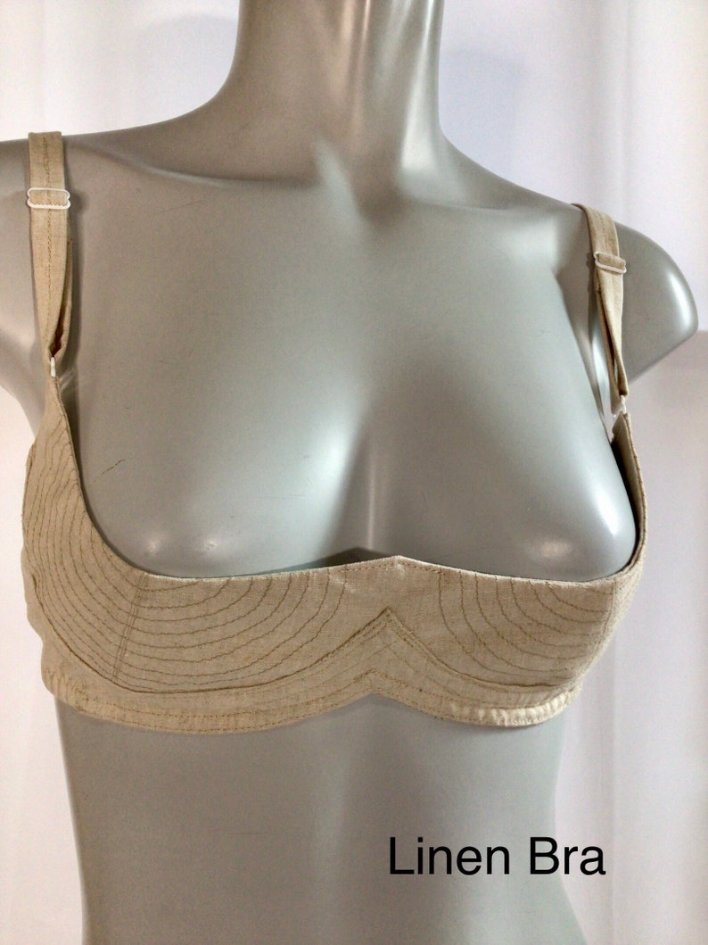 May include: A beige linen bra with adjustable straps. The bra has a unique design with a curved, scalloped neckline and a decorative stitch pattern. The text "Linen Bra" is printed below the bra.