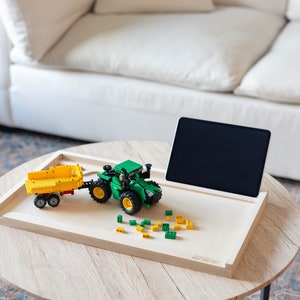 May include: A wooden tray with a green toy tractor and a yellow trailer, a tablet computer, and green and yellow toy building blocks on a light wood table.
