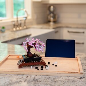May include: A wooden tray with a Lego bonsai tree, a tablet, and small black and white Lego pieces. The tray is labeled "BrickTray".