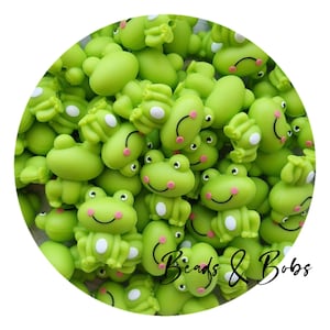 May include: A pile of small, lime green frog-shaped beads. Each frog has white eyes, a pink mouth, and a smiling expression. The text "Beads & Bobs" is written in a cursive font across the bottom of the image.