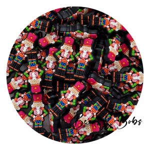 May include: A collection of colourful nutcracker charms. Each charm features a nutcracker soldier in a red hat and coat, with green and blue accents. The charms are black and have a glossy finish.