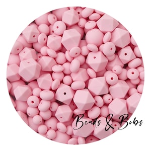 Könnte beinhalten: Eine Ansammlung rosafarbener Silikonperlen in verschiedenen Formen, darunter runde, ovale und geometrische Designs. Das Bild zeigt den Text "Beads & Bobs" in Schreibschrift, was auf ein Bastelzubehör oder Schmuckherstellungsthema hindeutet.