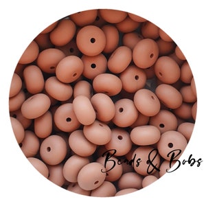 May include: A close-up image of a large quantity of light brown silicone beads. The beads are round with a hole in the center. The beads are arranged in a random pattern.