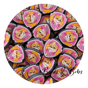 May include: A pile of pink, yellow, and black shoe charms, each depicting a cartoon dog character. The charms are shield-shaped with a pink border and a smiling dog face. The background is white.