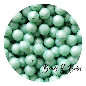 Può includere: Un insieme di perline rotonde verde chiaro con finitura perlata. Le perline sono sparse, riempiendo l'inquadratura. Il testo "Beads & Bobs" è scritto in corsivo in basso a destra.