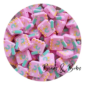 May include: A collection of pink silicone beads shaped like notebooks and pencils. The notebooks have yellow scalloped edges, ABC letters, and star details. The pencils are teal. The text "Beads & Bobs" is in the lower right corner.