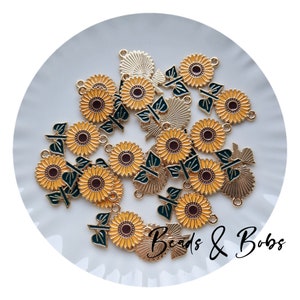 May include: A collection of sunflower charms with yellow petals, brown centers, and green leaves. The charms are gold-toned metal with enamel details. The text "Beads & Bobs" is written in a script font.