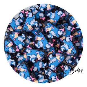 Könnte beinhalten: Eine Sammlung von blauen, grauen und rosa Eeyore-Charms. Jeder Charm zeigt den Esel aus Winnie Puuh mit einer rosa Schleife. Die Charms haben schwarze Ränder.