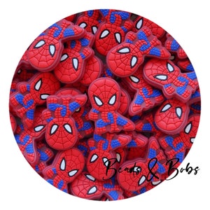 Op de afbeelding: Een verzameling kleine, rode en blauwe rubberen bedels met een spinnen-superheldontwerp, witte ogen en een zwart webpatroon. De tekst Beads & Bobs is in zwarte schrijfletters gedrukt in het rechtsonder gedeelte van de afbeelding.