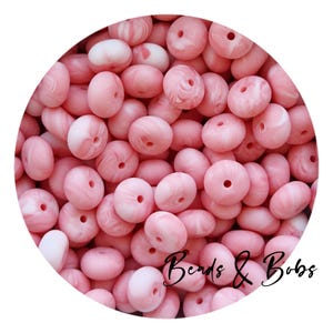 Pode incluir: Close-up de um monte de contas de silicone rosa com veios brancos. As contas têm um furo no centro e são projetadas para serem usadas na fabricação de joias ou como brinquedos para dentição.