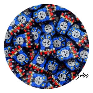 May include: A collection of blue, red, and yellow train-shaped charms. Each charm features a smiling face and is designed with details like wheels and a smokestack. The charms are arranged in a circular pattern.
