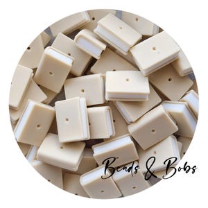 以下が含まれることがあります： 白いページが付いた、小さなベージュ色の本型のビーズのコレクション。各ビーズの表紙には小さな穴があります。「Beads & Bobs」の文字が筆記体で書かれています。