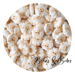 Può includere: Una collezione di abbellimenti bianchi a forma di pecora con facce e corpi color beige. Le pecore hanno lana soffice e sono disposte in un motivo circolare. Il testo "Beads & Bobs" è scritto in corsivo.