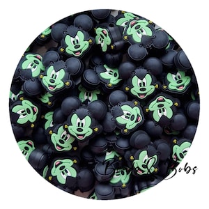 May include: A collection of black and light green rubber charms shaped like a cartoon mouse head with a stitched forehead detail. Each charm features a smiling face with a light green complexion, black ears, and a small black bolt on the side.