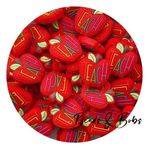 Pode incluir: Um close-up de um monte de pingentes vermelhos em forma de maçã com a palavra "TEACH" escrita em cores do arco-íris em cada pingente. Os pingentes são feitos de silicone e são perfeitos para adicionar um toque divertido e festivo às suas manualidades.