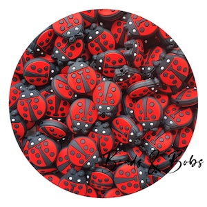 Op de afbeelding: Een verzameling rood-zwarte lieveheersbeestjesvormige bedels. Elke bedel heeft een rode body met zwarte stippen en een zwarte kop. De bedels zijn in een cirkelvormig patroon gerangschikt, met de woorden "Beads & Bobs" rechtsonder.