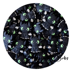 May include: A collection of black dragon-shaped charms with green eyes and white wings. The charms are arranged in a circle, with the word "Bobs" in cursive script in the lower right corner. These charms are likely for crafting or jewellery making.