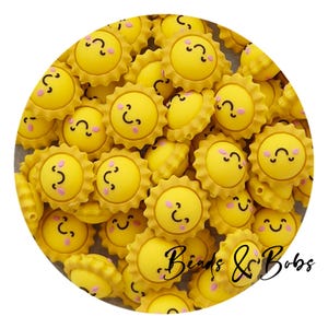 May include: A pile of yellow sun-shaped beads, each with a smiling face and pink cheeks. The beads have a scalloped edge, and the text "Beads & Bobs" is visible. These beads are suitable for crafting projects.