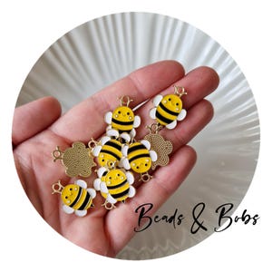 Pode incluir: Uma coleção de pingentes em forma de abelha com listras amarelas e pretas, asas brancas e rostos sorridentes. Os pingentes têm detalhes em metal dourado. O texto "Beads & Bobs" está escrito em itálico.