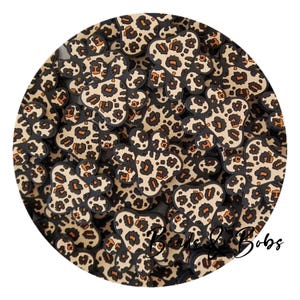 以下が含まれることがあります： ヒョウ柄の小さな茶色、黒、オレンジの動物の足跡の形をしたレジンチャームの山をクローズアップした画像です。