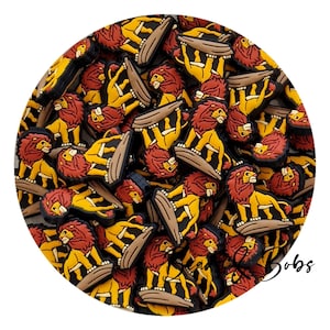 Op de afbeelding: Een verzameling kleine rubberen schoenbedels met een gele leeuw met donkerrode manen die op een bruine rots staat. Het ontwerp is meerdere keren in een cirkel gerangschikt. De tekst "Bead & Bobs" is zichtbaar in zwarte schrijfletters.