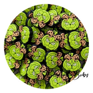 Può includere: Una collezione di ciondoli in silicone verde lime a forma di viso di personaggio con un fiocco color marrone chiaro a pois neri. Ogni ciondolo ha una base nera e un'espressione scolpita con occhi definiti e un naso piccolo. Il testo Beads & Bobs è visibile.