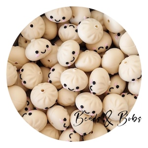 Pode incluir: Uma coleção de contas de silicone na cor creme em formato de pequenos pães cozidos no vapor, cada uma com um rosto sorridente preto estampado com bochechas rosadas e um orifício central para enfiar. O texto Beads & Bobs é visível no canto inferior direito.
