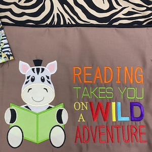 May include: A brown fabric bag with a zebra print design. The bag has a pocket with a zebra print fabric. The bag has an embroidered image of a zebra reading a book with the text "Reading takes you on a wild adventure".