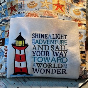 May include: A white pillow with a lighthouse design and the text "Shine a Light on Adventure and Sail Your Way Toward a World of Wonder". The pillow is on a gray cutting mat.