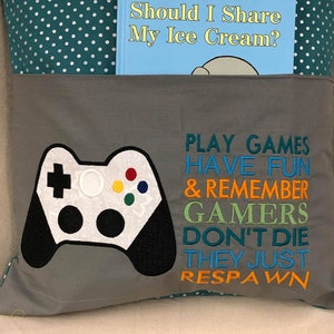 May include: A grey pillow with a pocket on the front. The pocket contains a book titled "Should I Share My Ice Cream?" The pillow has a white and black video game controller embroidered on the front with the text "PLAY GAMES HAVE FUN & REMEMBER GAMERS DON'T DIE THEY JUST RESPAWN" in orange, green, and blue.
