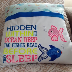 May include: White pillow with embroidered text "HIDDEN WITHIN THE OCEAN DEEP THE FISHES READ BEFORE THEY SLEEP" and two embroidered fish, one pink with white polka dots and one blue with a white pattern.