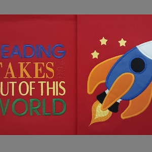 Könnte beinhalten: Rotes Stoffbuch mit dem Text "Reading Takes You Out of This World" in bunten Buchstaben auf der linken Seite und einer blauen und orangen Rakete mit weißem Oberteil und gelben Flammen auf der rechten Seite. Auf der rechten Seite des Buches befinden sich fünf gelbe Sterne.