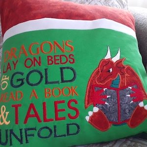 May include: Green pillow with embroidered text that reads "Dragons lay on beds of gold read a book & tales unfold". A red dragon with wings is embroidered on the pillow, holding a book.