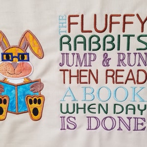 Może przedstawiać: Haftowany wzór przedstawiający brązowego królika w okularach czytającego niebieską książkę. Tekst brzmi "The Fluffy Rabbits Jump & Run Then Read A Book When Day Is Done."
