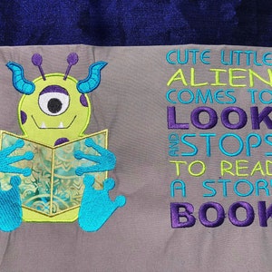 May include: A gray and purple embroidered design of a green alien reading a book with the text "Cute little alien comes to look and stops to read a story book".