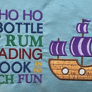 May include: A blue fabric background with the text "Yo ho ho and a bottle of rum reading a book is so much fun" in colorful embroidery. A brown embroidered ship with purple sails is on the right side of the image.