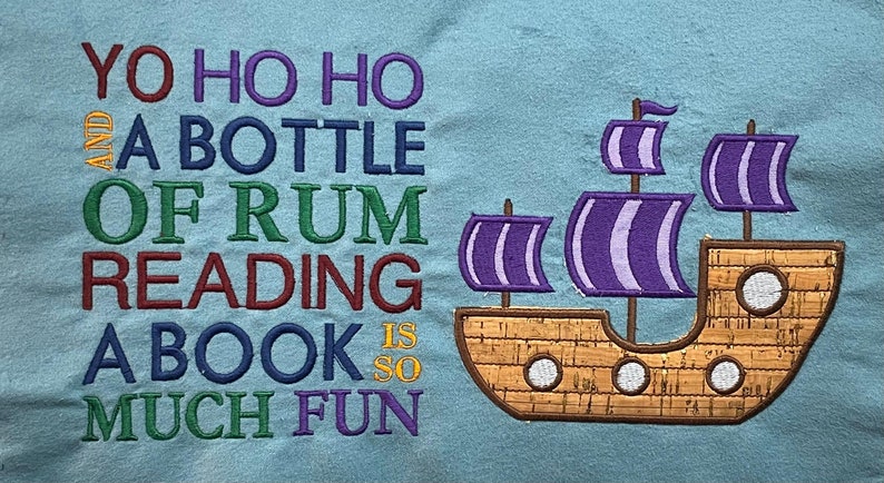 May include: A blue fabric background with the text "Yo ho ho and a bottle of rum reading a book is so much fun" in colorful embroidery. A brown embroidered ship with purple sails is on the right side of the image.