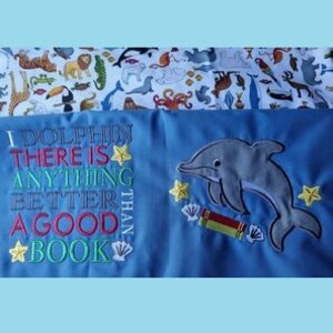 May include: A blue fabric panel with embroidered text and a dolphin design. The text reads "I DOLPHIN THERE IS ANYTHING BETTER THAN A GOOD BOOK." The dolphin is gray, and there are yellow star accents.