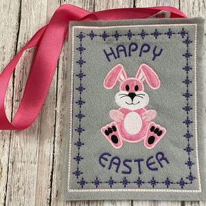 May include: A grey felt Easter bag with a pink bunny and the text "Happy Easter" embroidered on it. The bag has a pink ribbon attached to it.