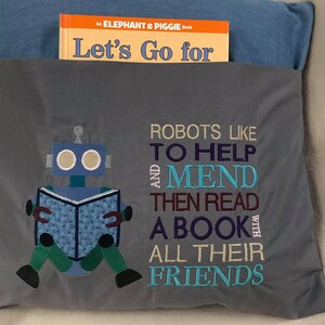 May include: Gray pillow with embroidered text that reads "Robots like to help and mend then read a book with all their friends". A blue robot with a book is embroidered in the center of the pillow. The pillow is sitting on a blue and gray surface.