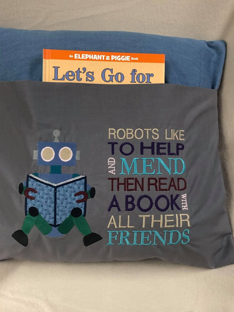May include: Gray pillow with embroidered text that reads "Robots like to help and mend then read a book with all their friends". A blue robot with a book is embroidered in the center of the pillow. The pillow is sitting on a blue and gray surface.
