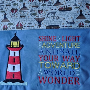 May include: A blue fabric with a red and white lighthouse embroidered on the left side. The text "Shine a light on adventure and sail your way toward a world of wonder" is embroidered in red, yellow, and blue on the right side.