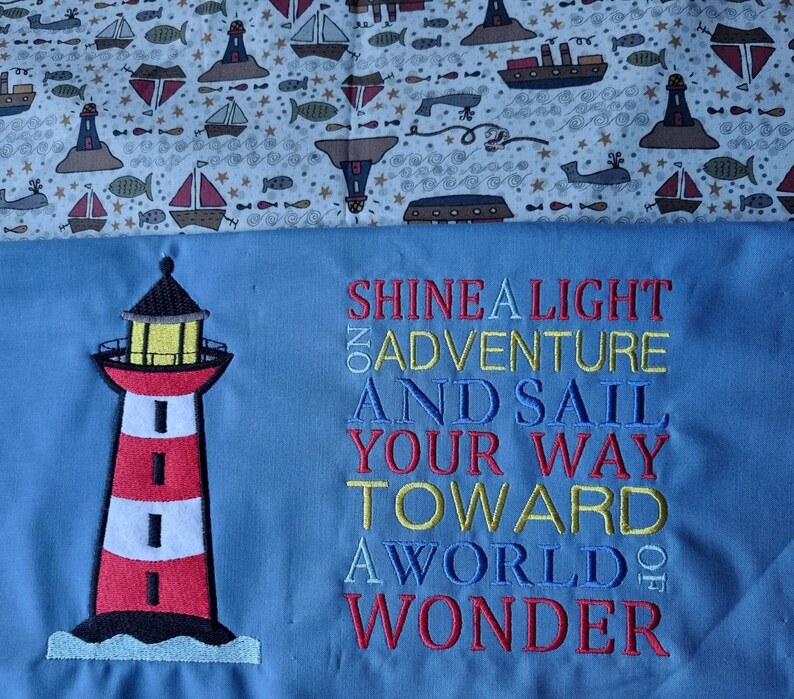 May include: A blue fabric with a red and white lighthouse embroidered on the left side. The text "Shine a light on adventure and sail your way toward a world of wonder" is embroidered in red, yellow, and blue on the right side.