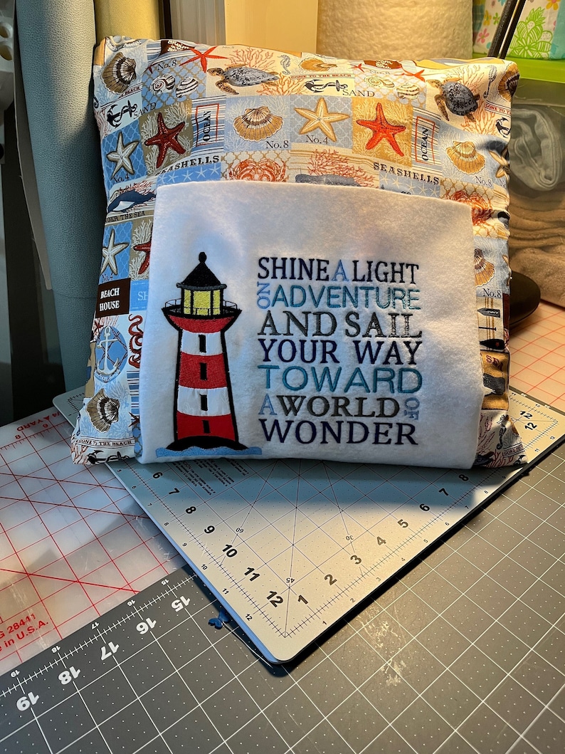 May include: A white pillow with a lighthouse design and the text "Shine a Light on Adventure and Sail Your Way Toward a World of Wonder". The pillow is on a gray cutting mat.