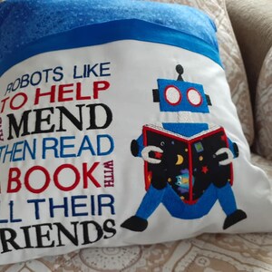 May include: White pillow with blue trim featuring an embroidered robot reading a book. The text reads "Robots like to help and mend then read a book with all their friends".