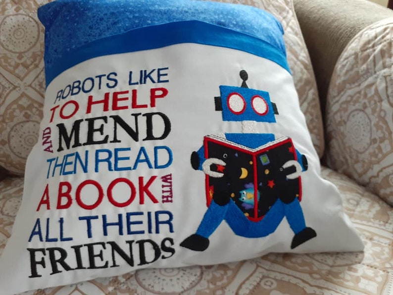 May include: White pillow with blue trim featuring an embroidered robot reading a book. The text reads "Robots like to help and mend then read a book with all their friends".