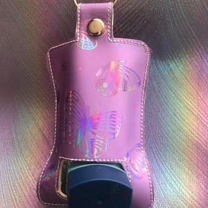 May include: A pink leather keychain holder with a gold ring and a snap closure. The holder is decorated with iridescent butterfly designs and is designed to hold a small bottle of hand sanitizer.