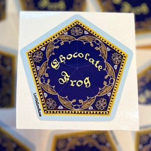 May include: A blue and gold pentagon-shaped sticker with the words "Chocolate Frog" in a gold script font. The sticker has a decorative border and a small white background.