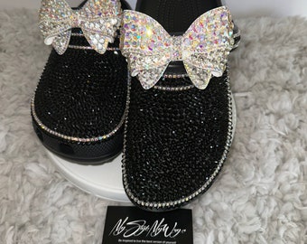 Puede incluir: Zuecos negros con un acabado negro brillante y dos grandes lazos de cristal iridiscente en la parte superior. Los zuecos están sobre una superficie blanca y esponjosa con una tarjeta de visita negra que dice "My Style My Way" y "Inspírate para vivir la mejor versión de ti mismo."